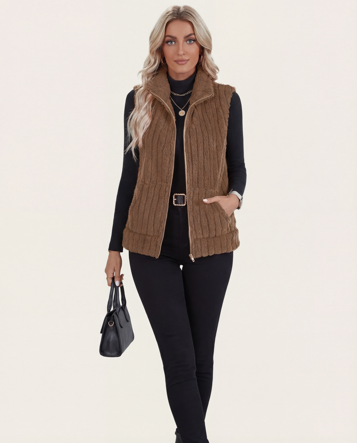 Woman wearing a brown knit vest over a black top and pants, holding a black handbag on a beige background.